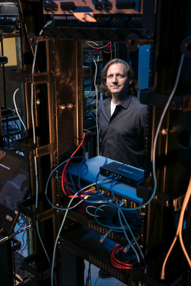 Jim Jones wears a dark shirt and stands in an IT database lab with wires and boxes.