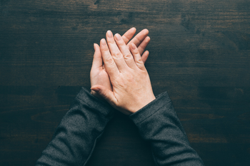 A person's two hands are placed together, right over left, on a wooden table 